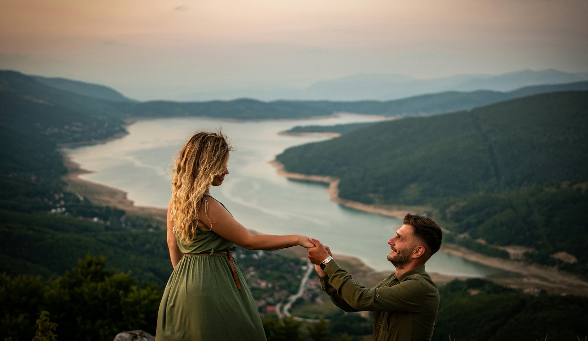 Man proposing to a woman on a mountain overlooking a winding river at sunset, creating a romantic moment with scenic natural views.