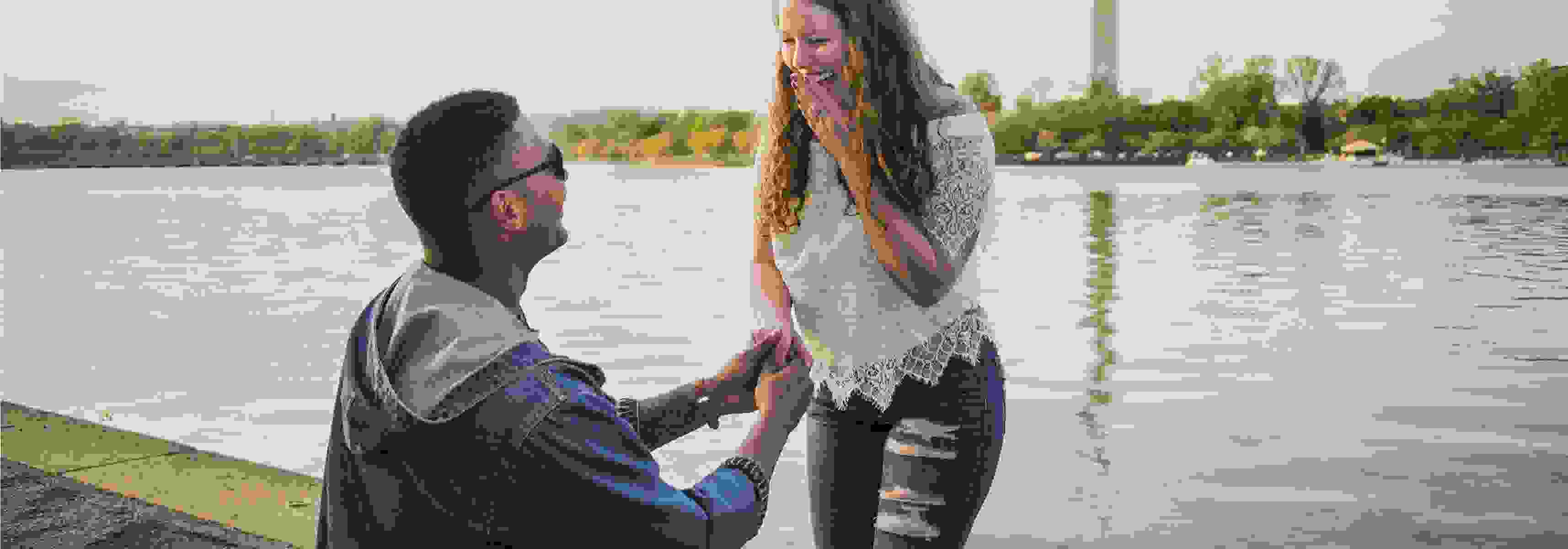 Man proposing to a smiling woman near a body of water with the Washington Monument in the background