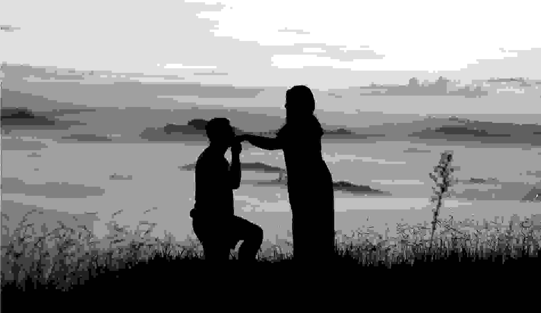 Silhouette of a man proposing to a woman on a hilltop at sunrise, with a misty valley and distant mountains in the background. 