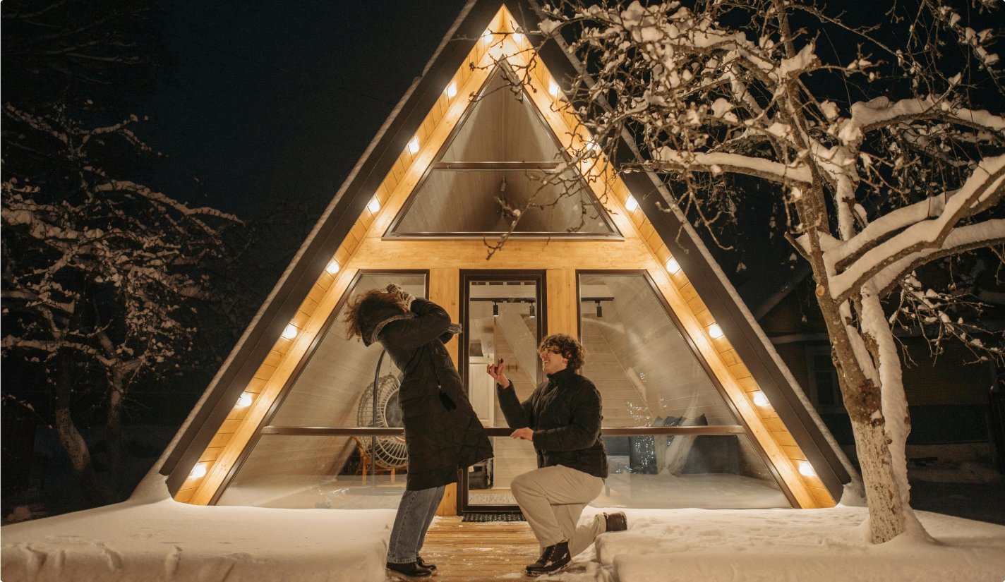 Man proposing to a surprised woman outside a warmly lit A-frame cabin at night, surrounded by snow-covered trees and winter scenery.