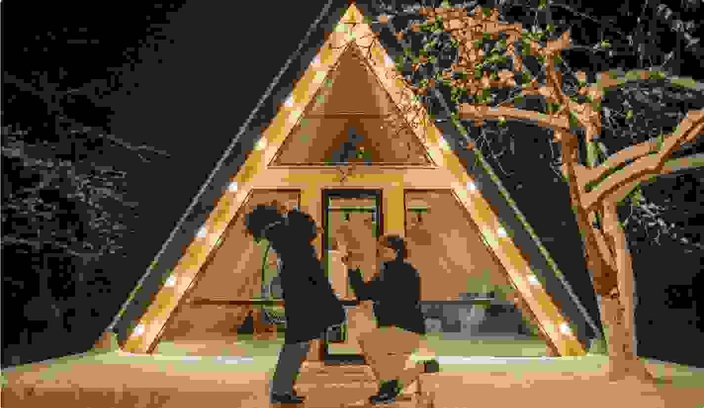 Man proposing to a surprised woman outside a warmly lit A-frame cabin at night, surrounded by snow-covered trees and winter scenery. 