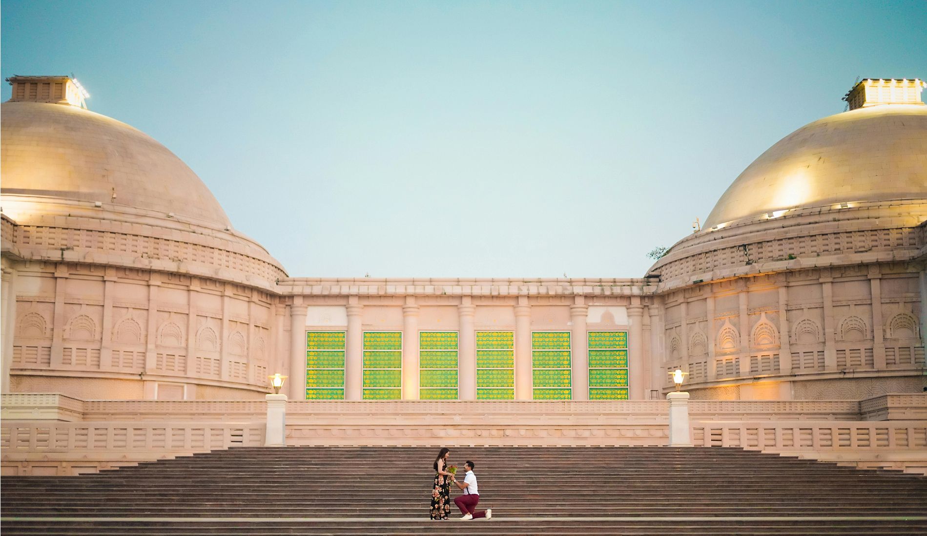 Man proposing to a woman on the steps of a grand architectural building with domed rooftops and vibrant green windows, during twilight.