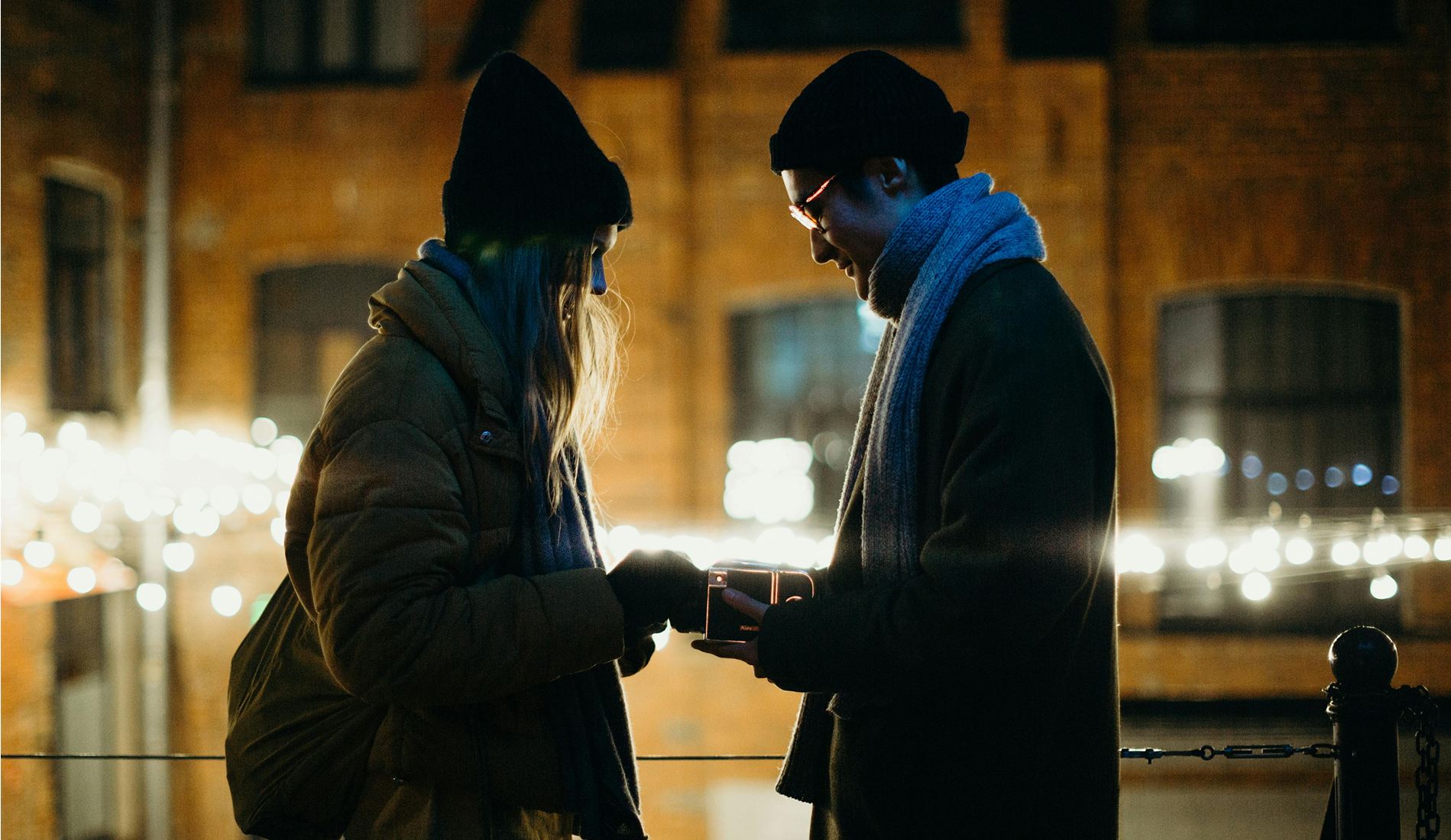 Couple standing close together on a winter evening, with the man holding an open ring box as string lights glow in the background, creating a romantic urban proposal setting.