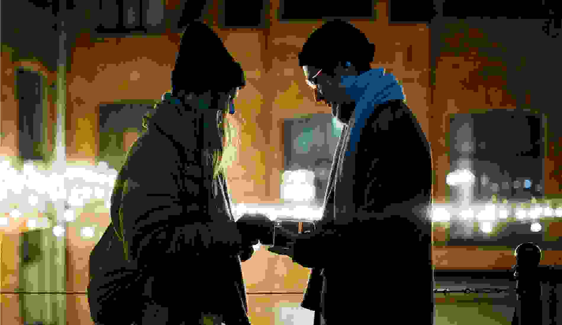 Couple standing close together on a winter evening, with the man holding an open ring box as string lights glow in the background, creating a romantic urban proposal setting. 