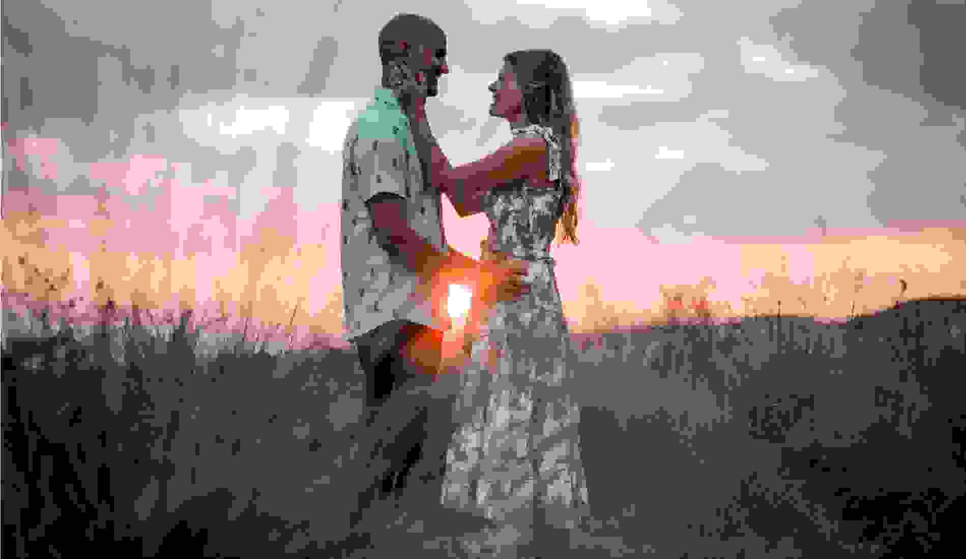 Couple standing close together in a field at sunset, smiling and holding each other lovingly with tall grass and a warm sky in the background.