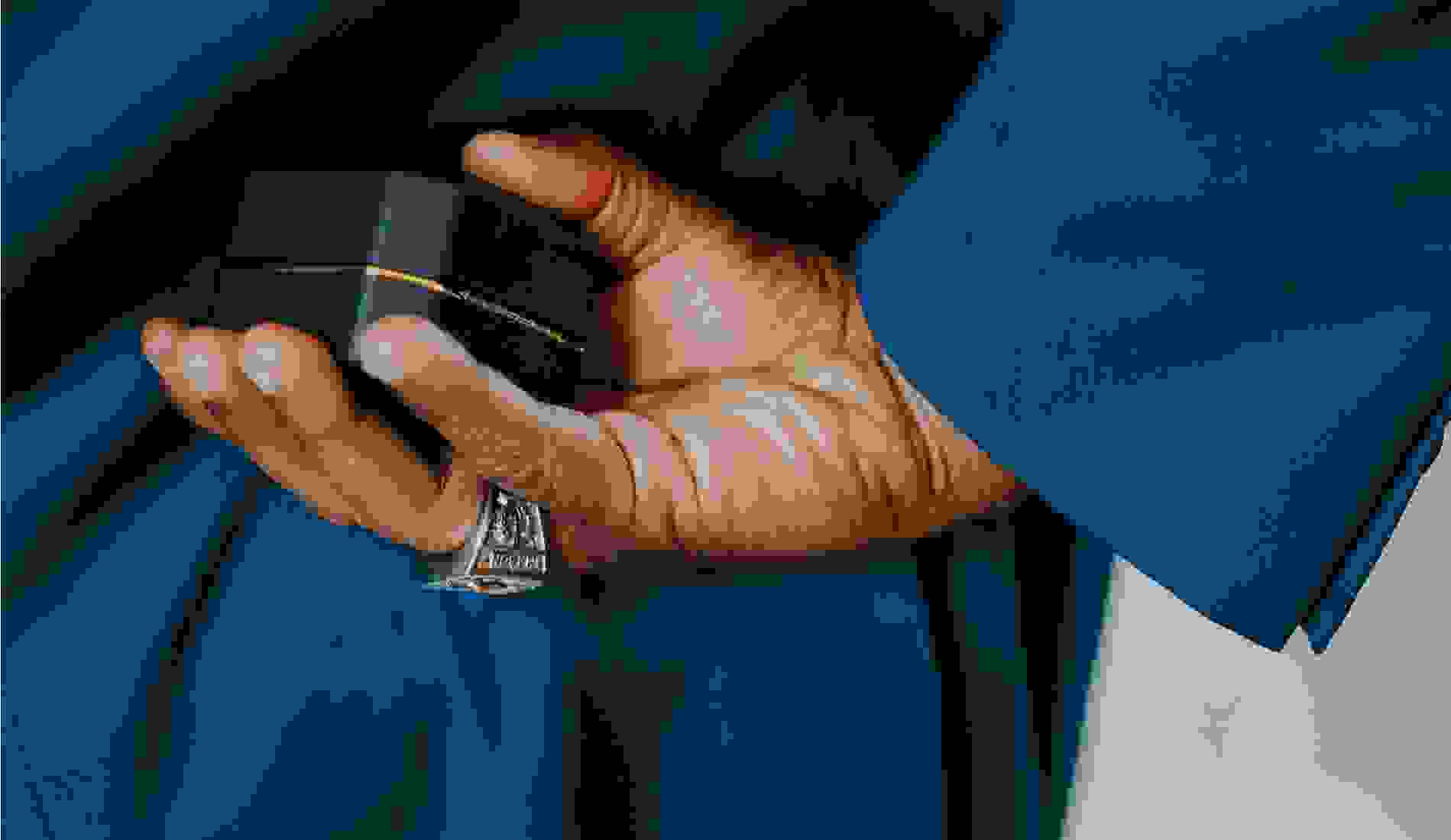 Close-up of a man’s hand holding a closed black ring box behind his back, dressed in a blue suit and wearing a statement ring.