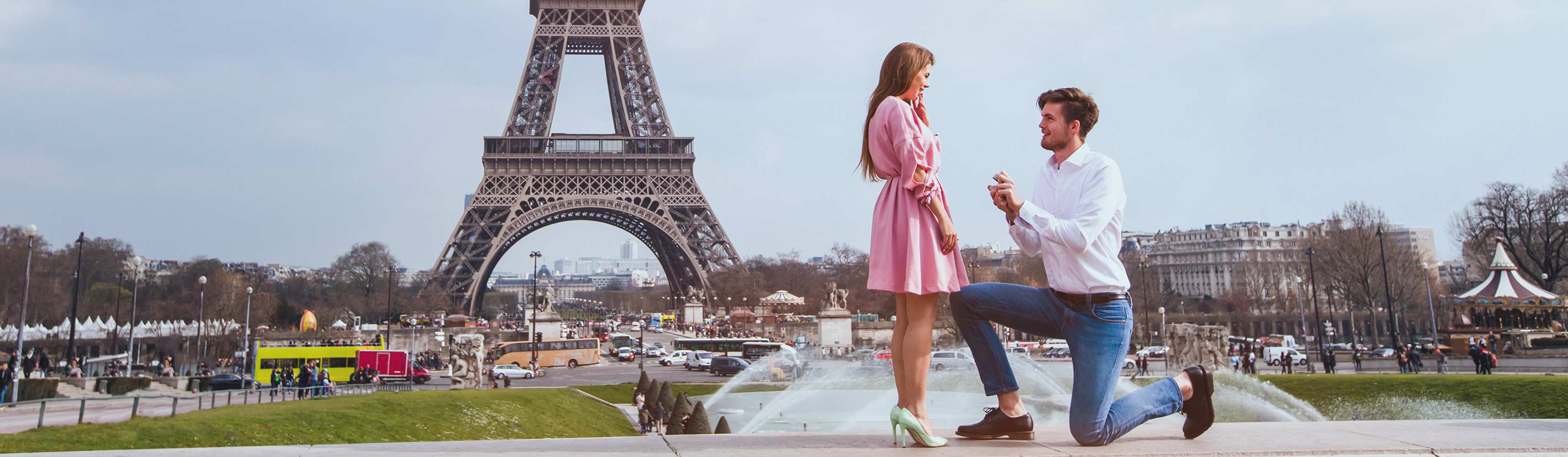 Man proposing to a woman in front of the Eiffel Tower in Paris, holding an open ring box while kneeling as she reacts with surprise and joy
