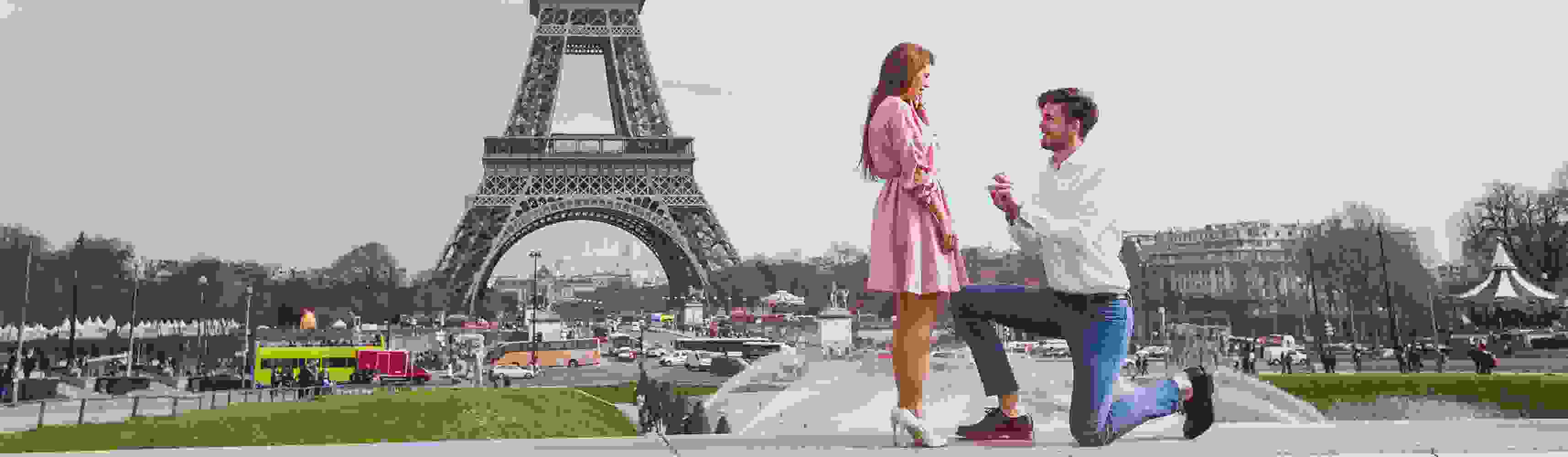 Man proposing to a woman in front of the Eiffel Tower in Paris, holding an open ring box while kneeling as she reacts with surprise and joy