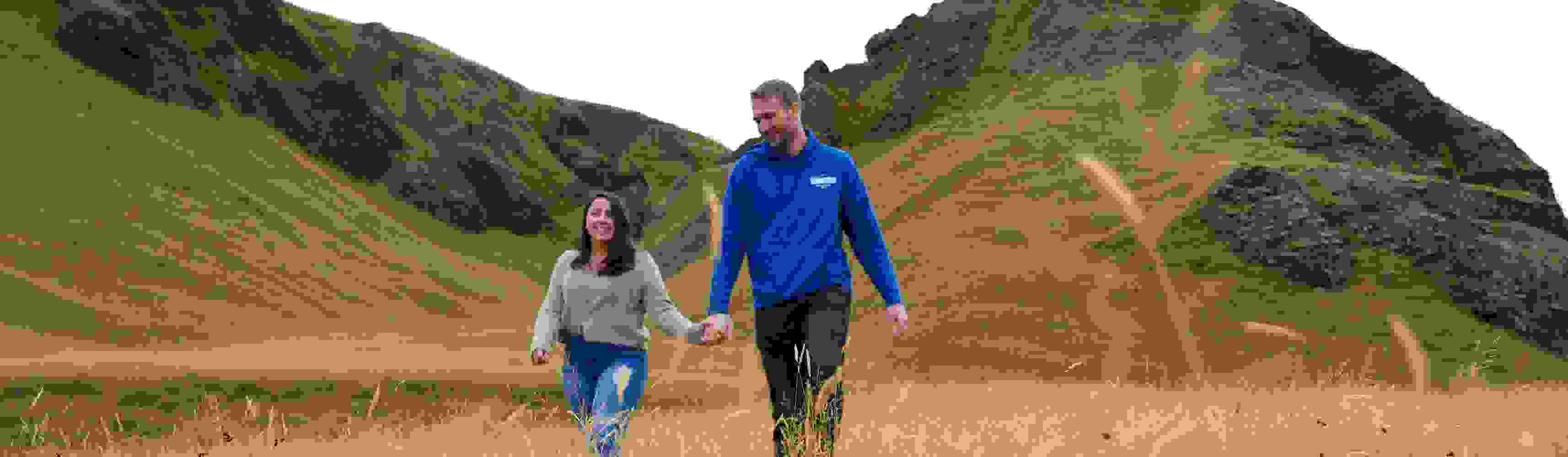 Couple holding hands and smiling while walking through a golden grassy field with rocky hills in the background, enjoying a romantic moment in nature
