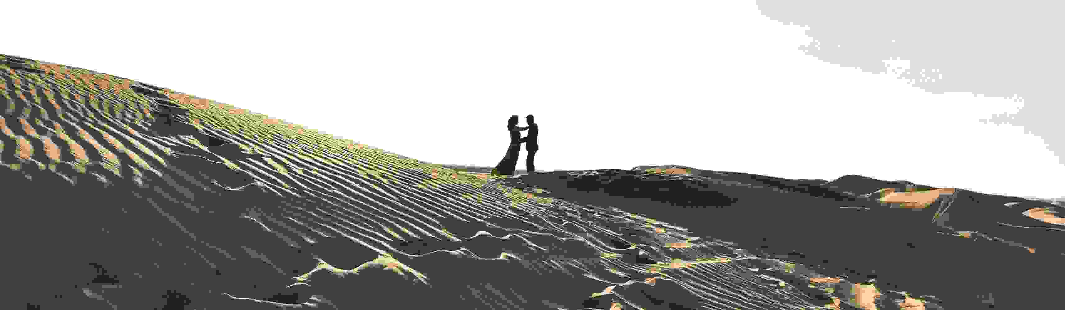 Couple holding hands and smiling while walking through a golden grassy field with rocky hills in the background, enjoying a romantic moment in nature