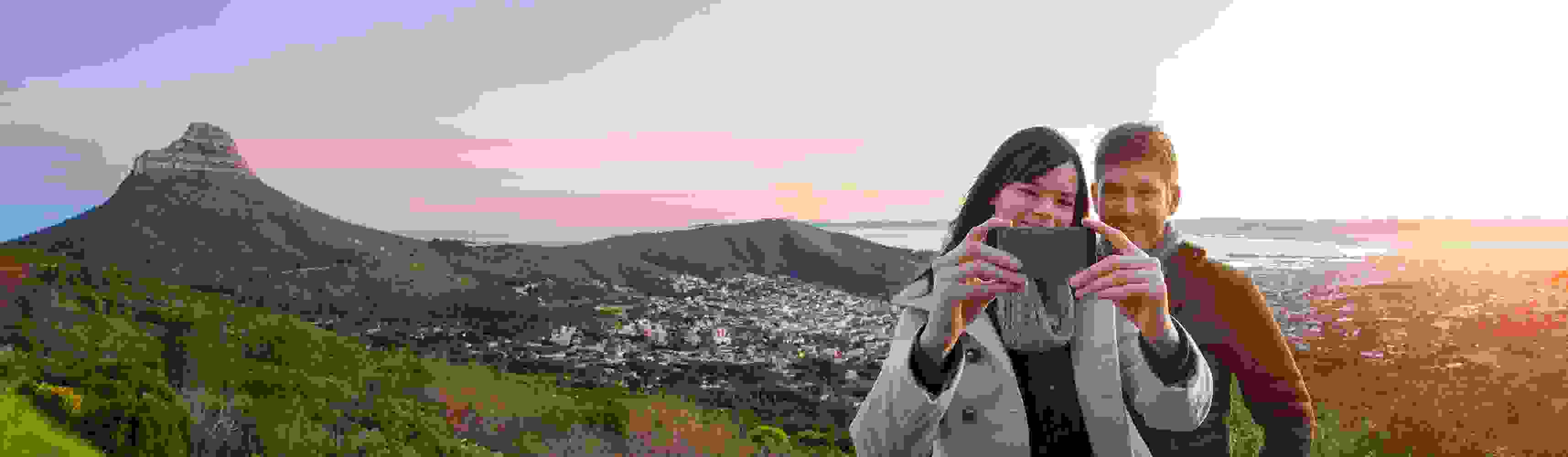 Smiling couple taking a selfie with a scenic mountain and city landscape behind them at sunset, capturing a joyful travel moment together