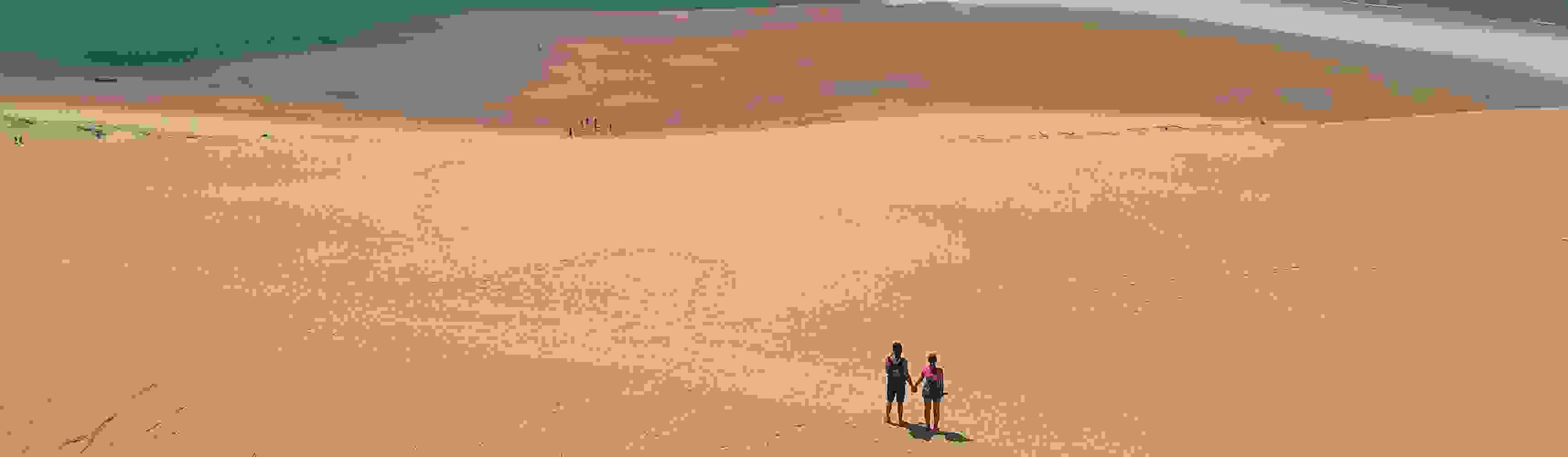 Couple holding hands and standing atop a vast sandy dune, overlooking a tidal shoreline with scattered footprints and distant water below
