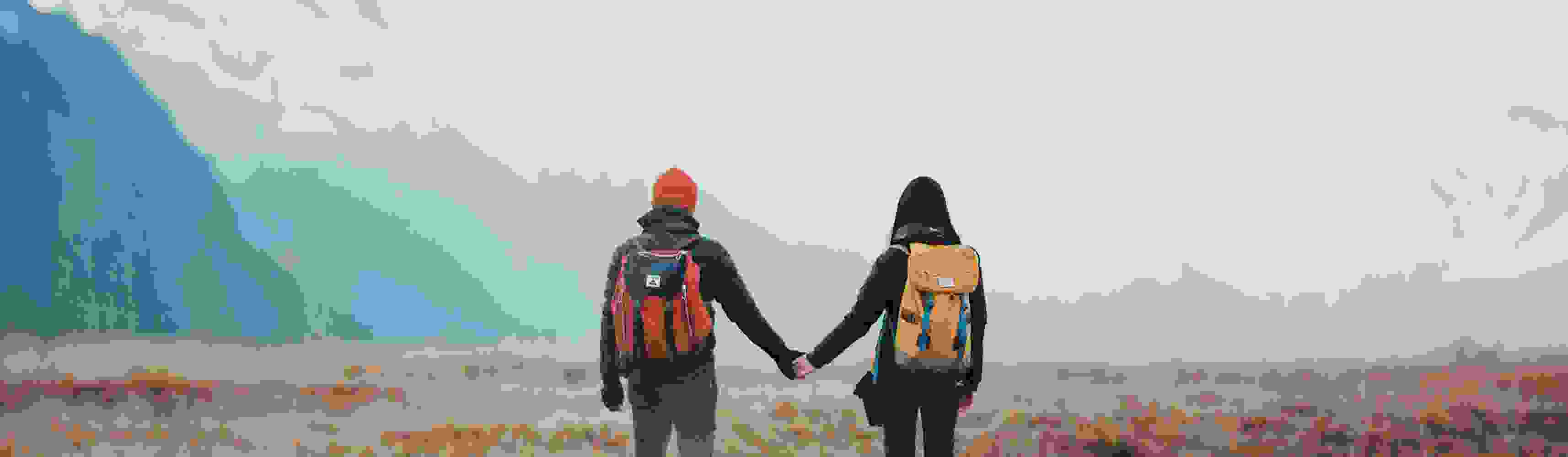 Couple holding hands and hiking through a vast alpine meadow with snow-capped mountains in the distance, each carrying a backpack and dressed for adventure
