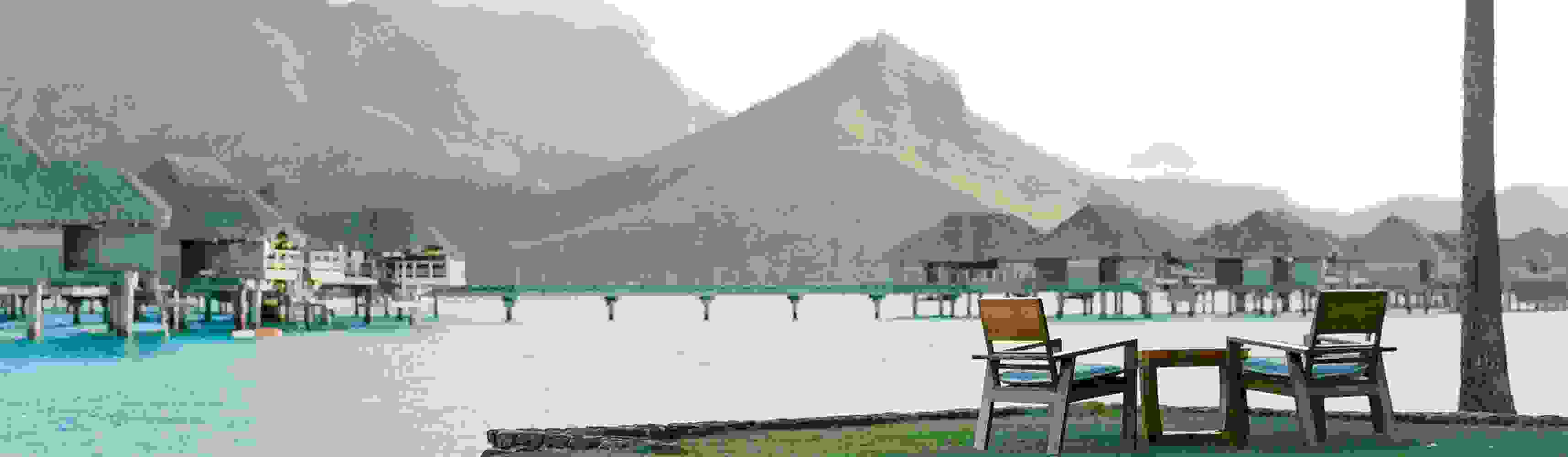Two empty wooden chairs and a small table set on a grassy edge overlooking turquoise water and overwater bungalows, with lush green mountains in the background