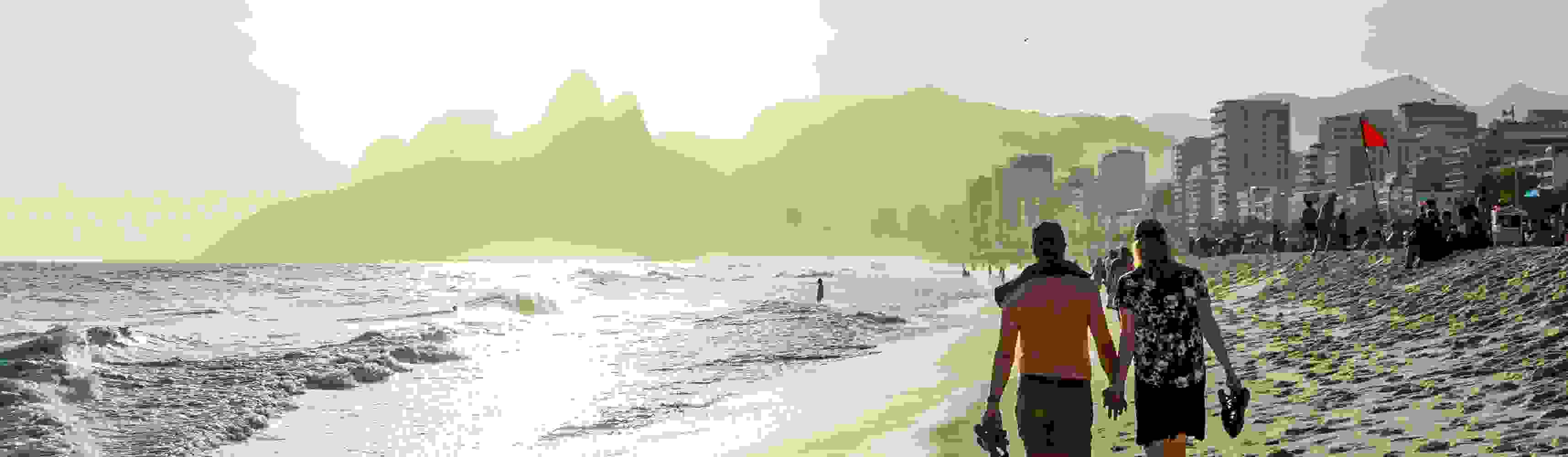 A couple holding hands walking along a bustling beach at sunset, with waves crashing on the shore and a city skyline and mountains in the hazy background