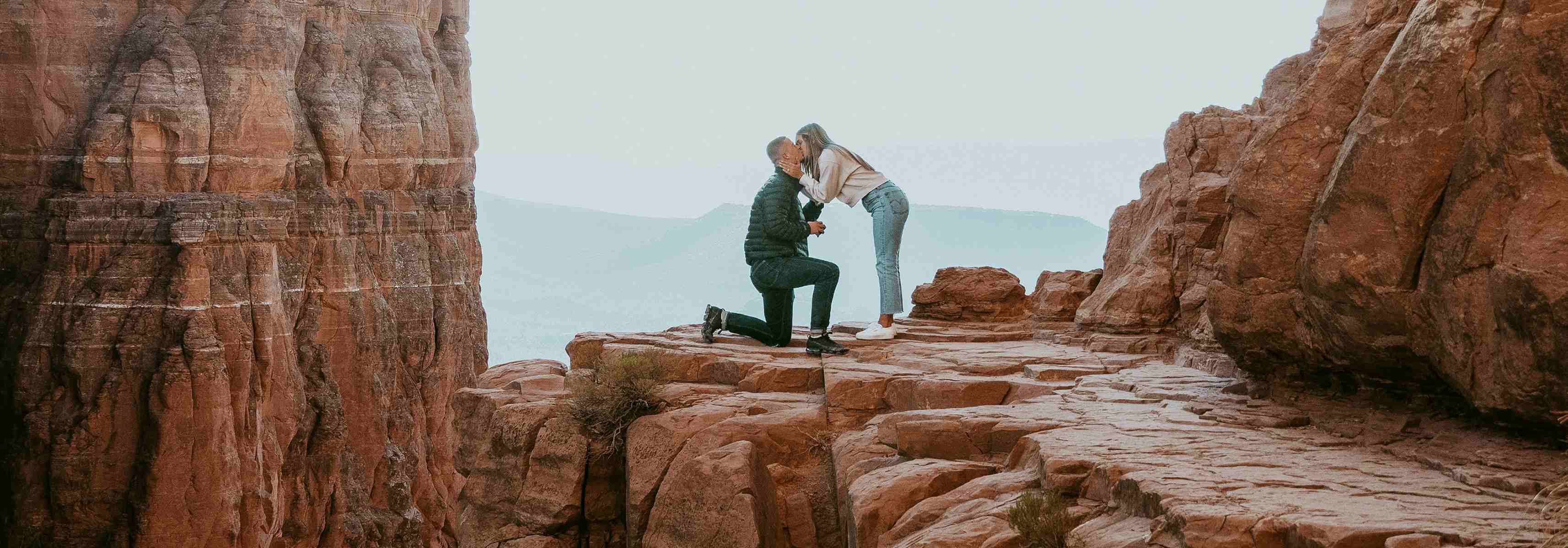 Man proposing to a woman on a scenic cliffside surrounded by red rock formations.