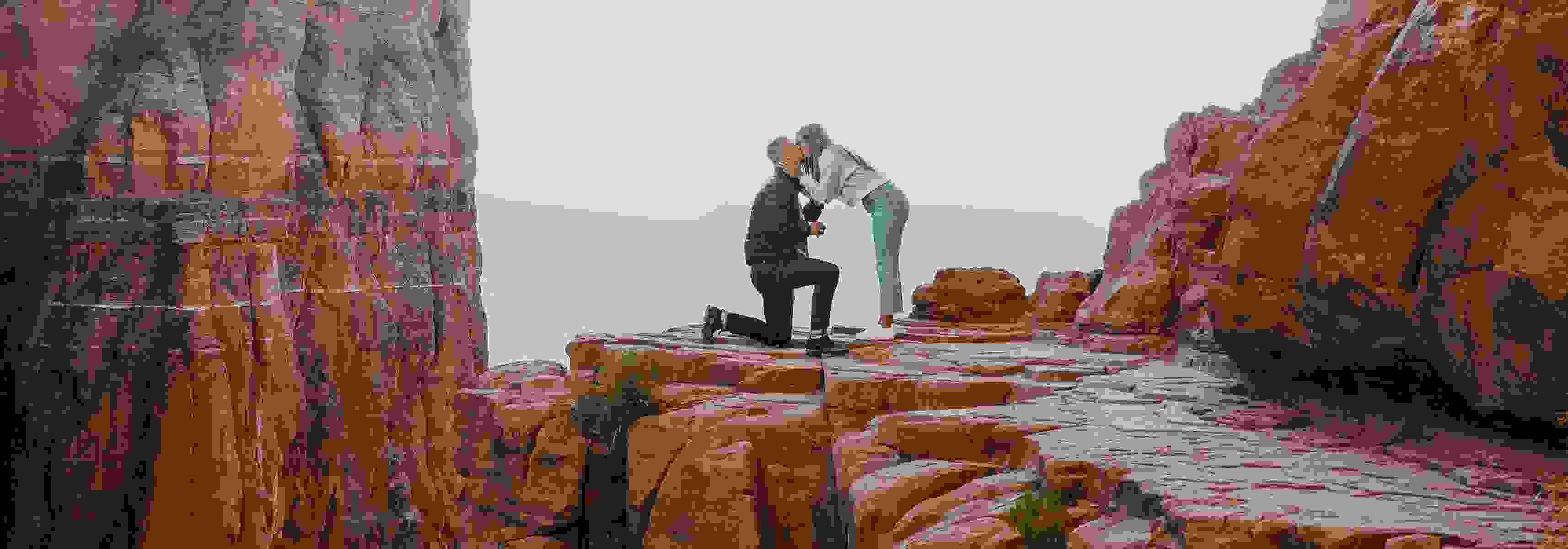 Man proposing to a woman on a scenic cliffside surrounded by red rock formations.