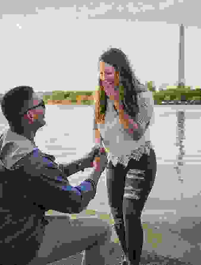 Man proposing to a smiling woman near a body of water with the Washington Monument in the background