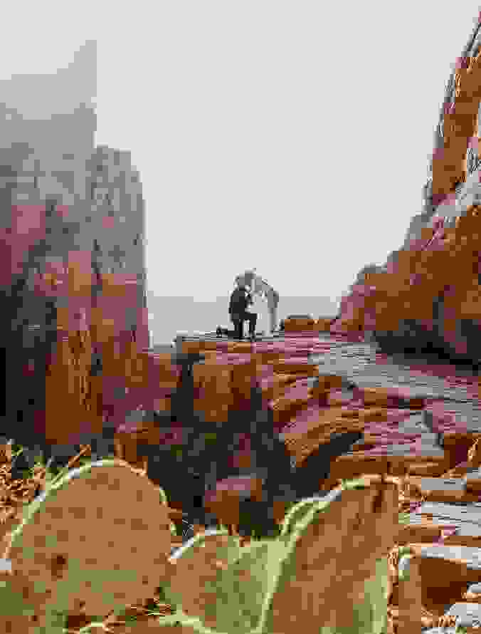 Man proposing to a woman on a scenic cliffside surrounded by red rock formations.