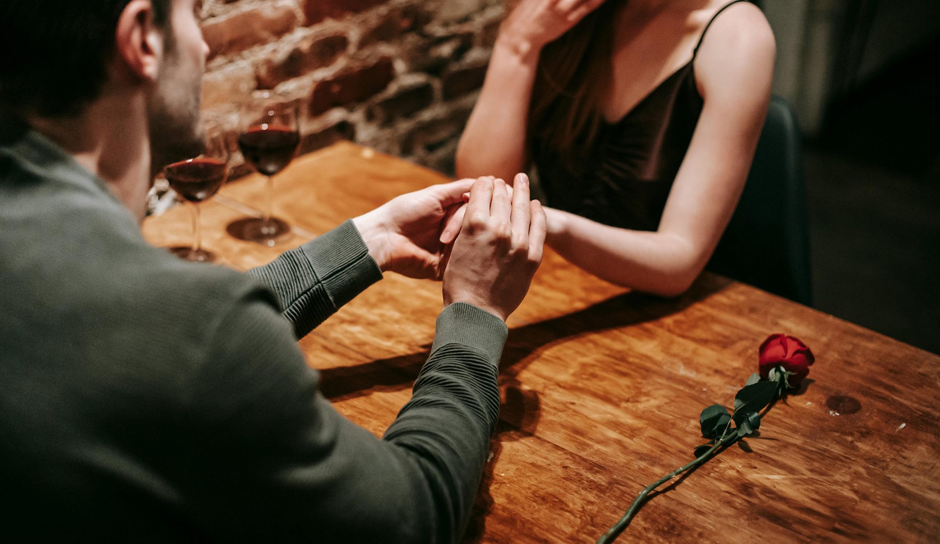 Man placing a ring on a woman's finger during a romantic dinner, with two glasses of red wine and a single red rose on the wooden table