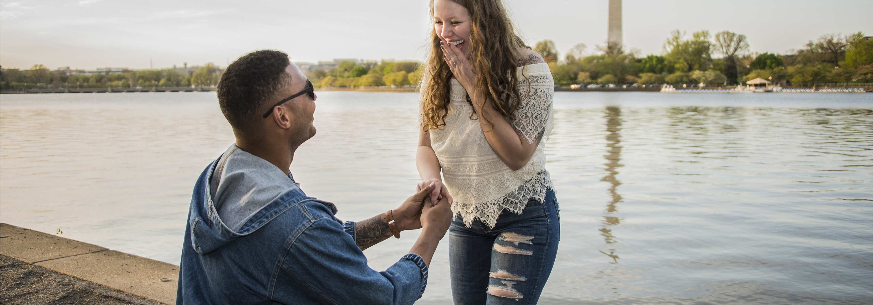 Man proposing to a smiling woman near a body of water with the Washington Monument in the background