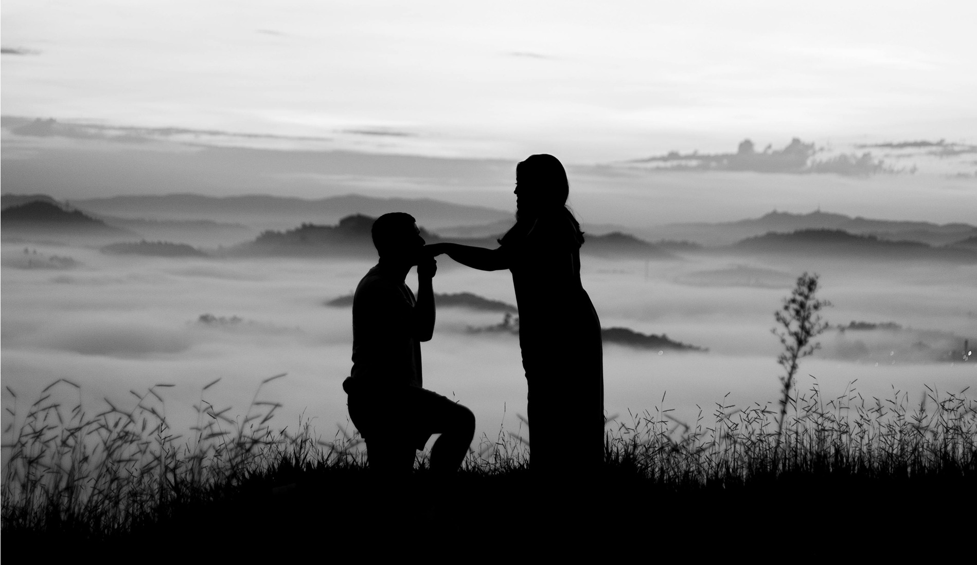 Silhouette of a man proposing to a woman on a hilltop at sunrise, with a misty valley and distant mountains in the background. 