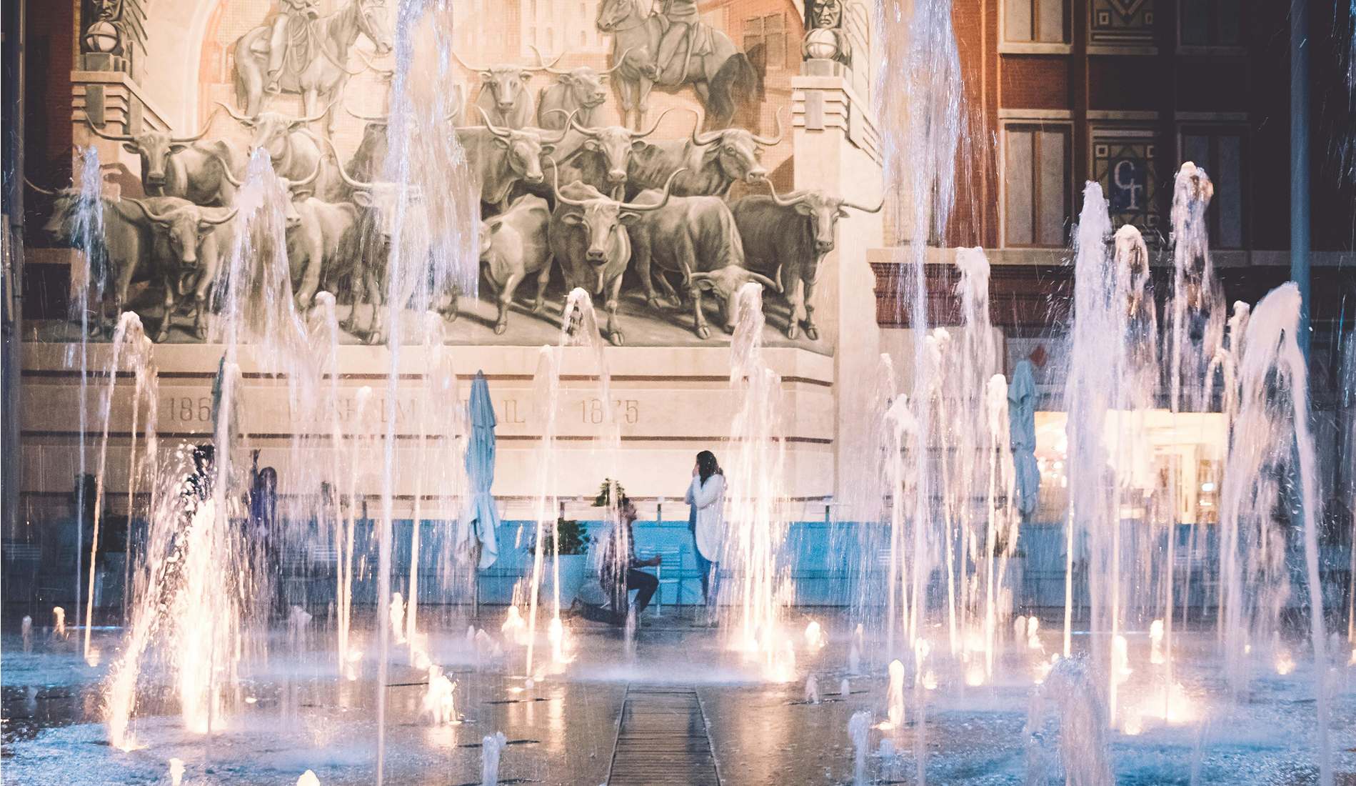 Man proposing to a woman amid illuminated water fountains in front of a large mural depicting cattle and cowboys, capturing a romantic moment in a public square. 