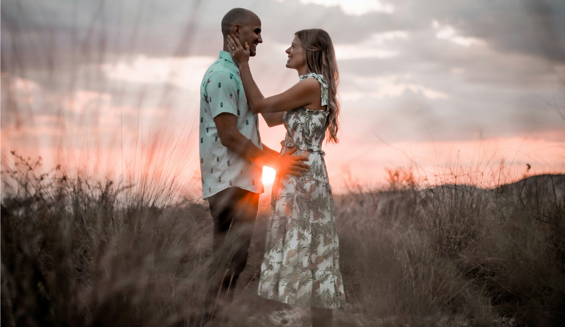 Couple standing close together in a field at sunset, smiling and holding each other lovingly with tall grass and a warm sky in the background.