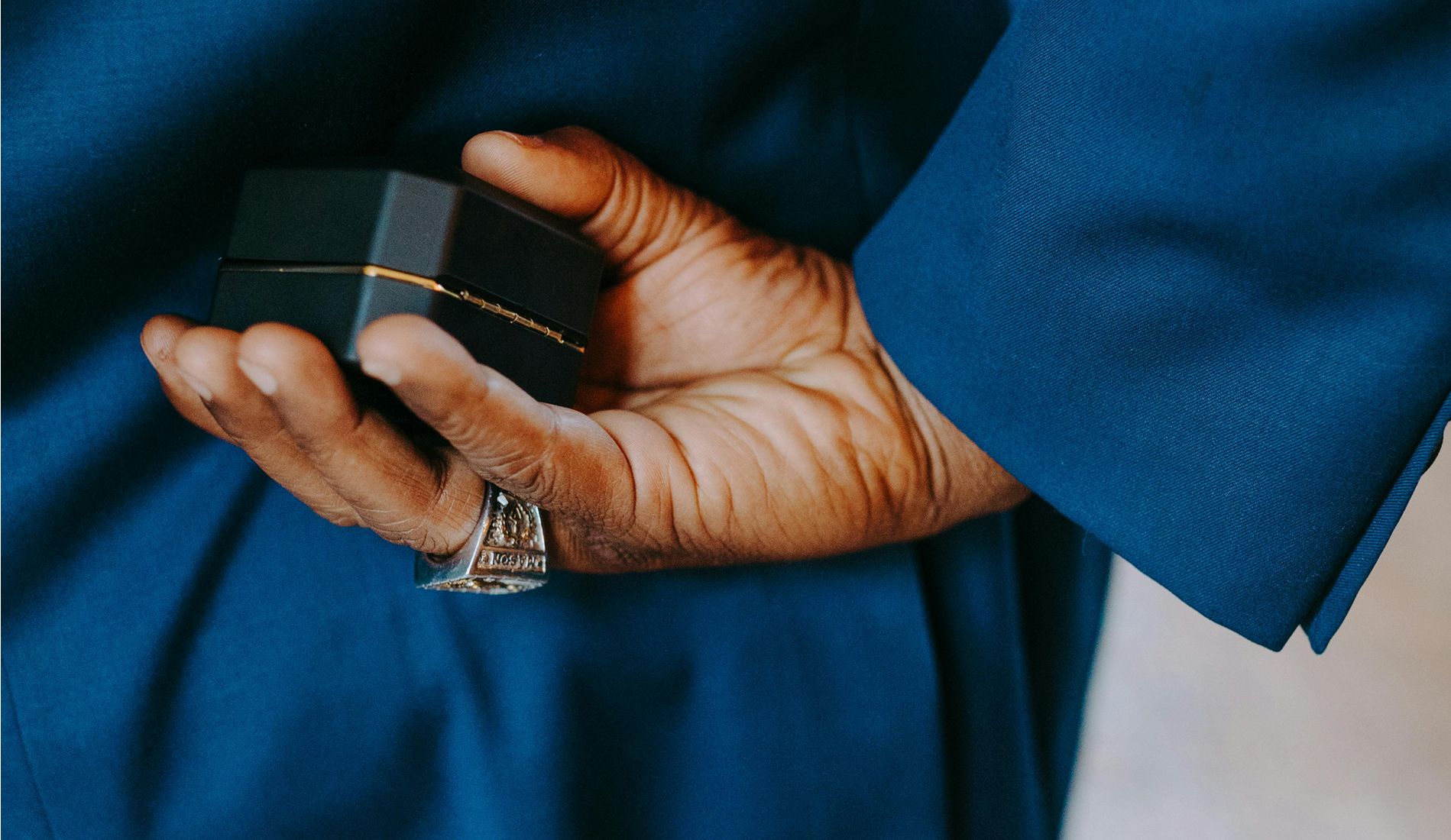 Close-up of a man’s hand holding a closed black ring box behind his back, dressed in a blue suit and wearing a statement ring.