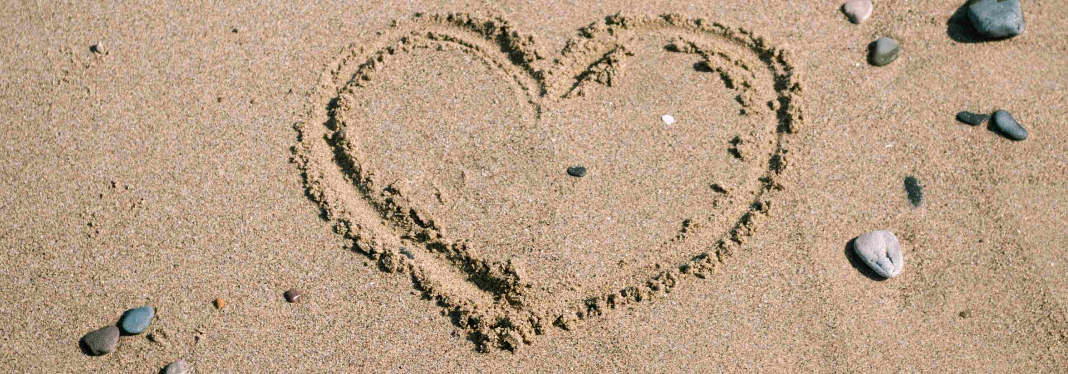 Heart shape drawn in the sand surrounded by scattered pebbles on a beach.