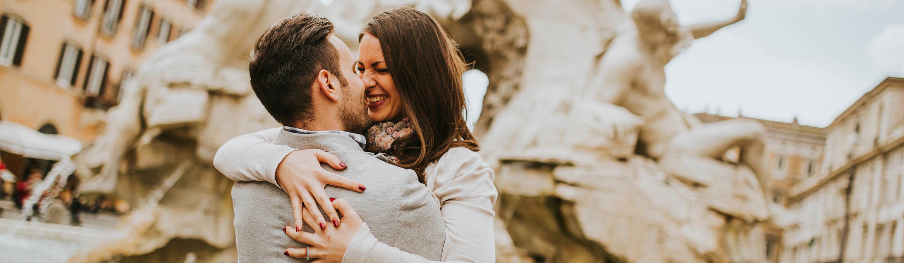 Happy couple embracing and smiling in front of a historic fountain in a European city, celebrating a romantic moment together