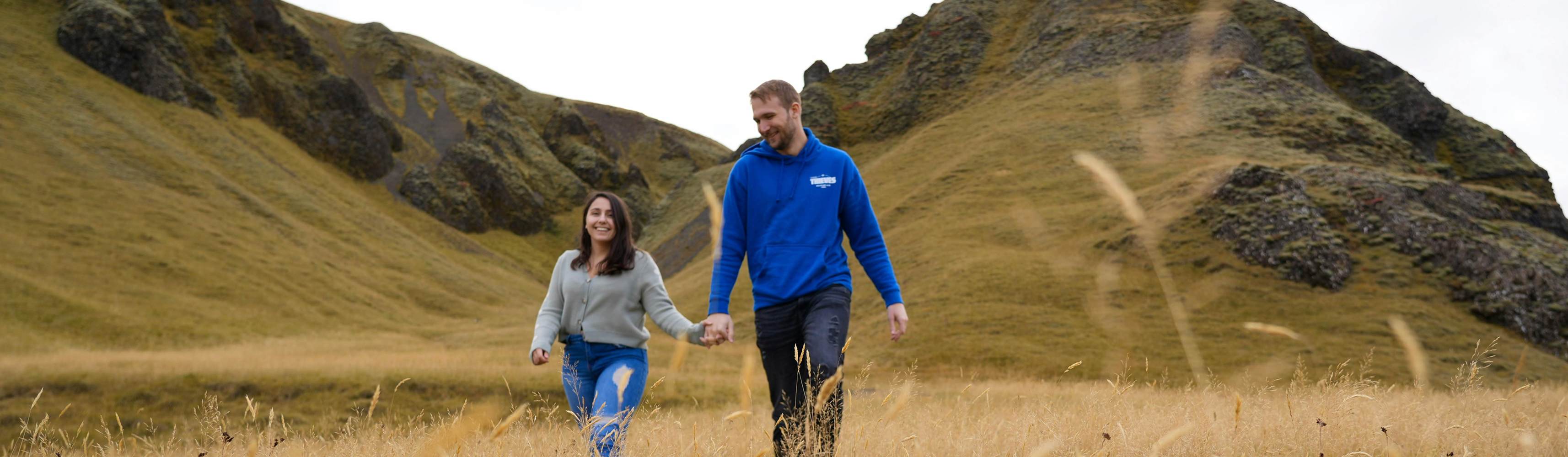 Couple holding hands and smiling while walking through a golden grassy field with rocky hills in the background, enjoying a romantic moment in nature
