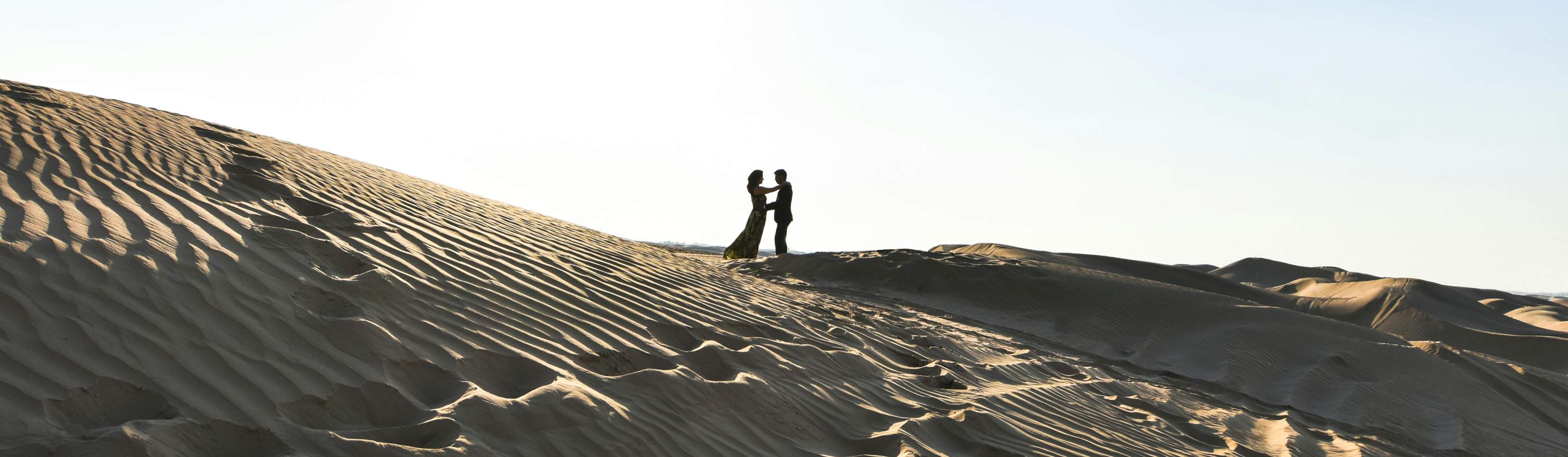 Couple holding hands and smiling while walking through a golden grassy field with rocky hills in the background, enjoying a romantic moment in nature