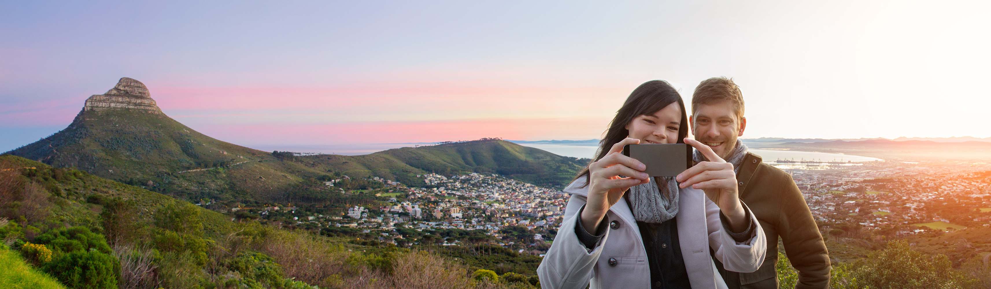 Smiling couple taking a selfie with a scenic mountain and city landscape behind them at sunset, capturing a joyful travel moment together
