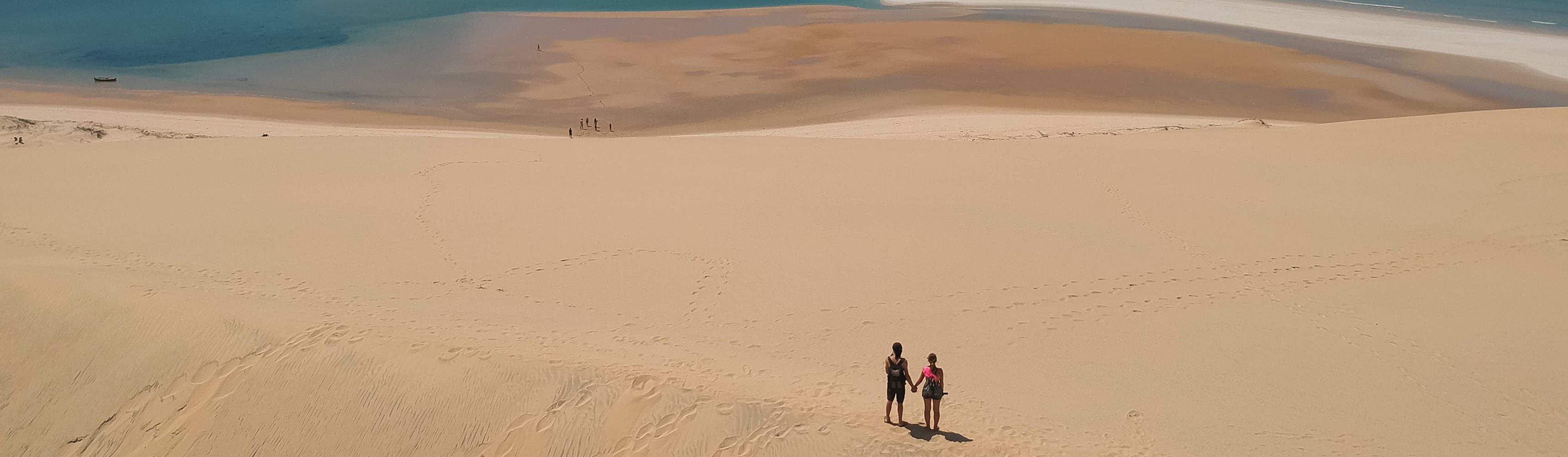 Couple holding hands and standing atop a vast sandy dune, overlooking a tidal shoreline with scattered footprints and distant water below