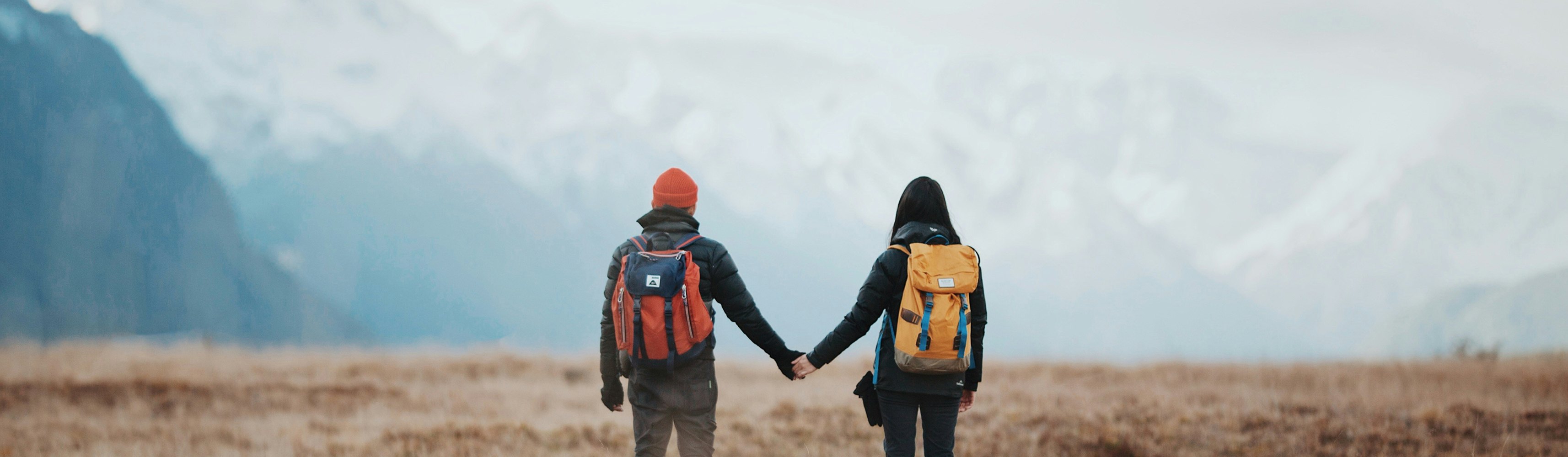 Couple holding hands and hiking through a vast alpine meadow with snow-capped mountains in the distance, each carrying a backpack and dressed for adventure