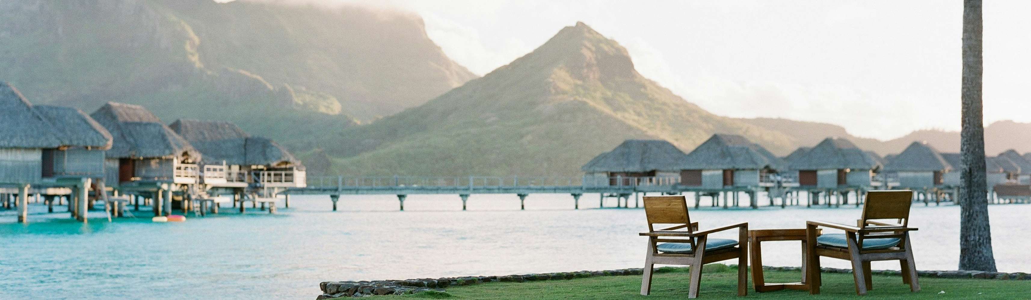 Two empty wooden chairs and a small table set on a grassy edge overlooking turquoise water and overwater bungalows, with lush green mountains in the background