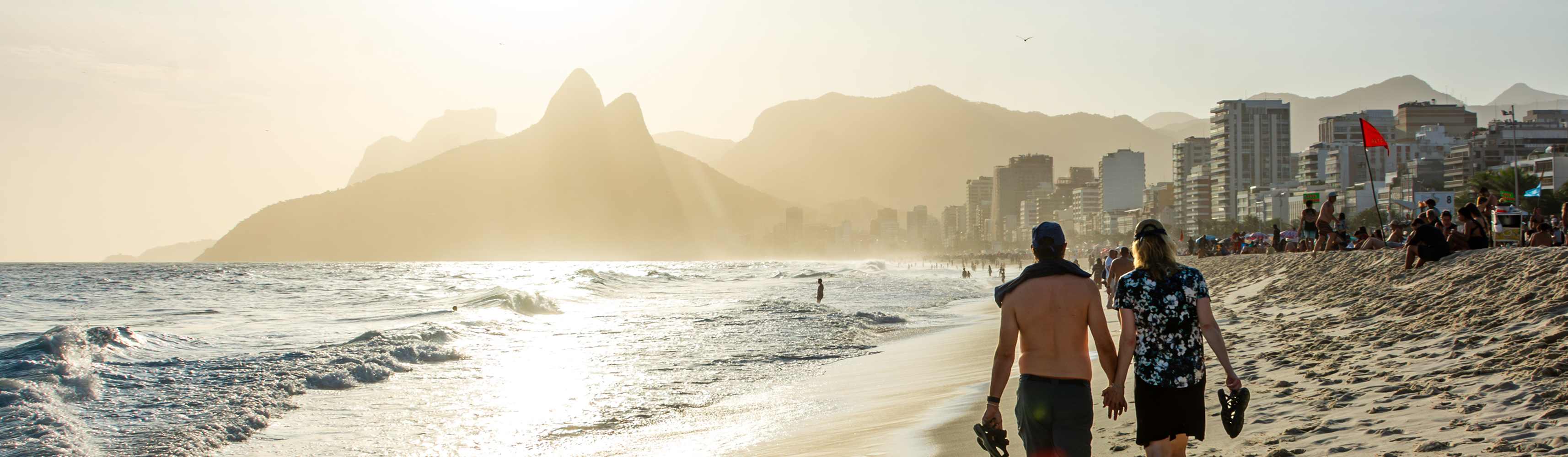 A couple holding hands walking along a bustling beach at sunset, with waves crashing on the shore and a city skyline and mountains in the hazy background