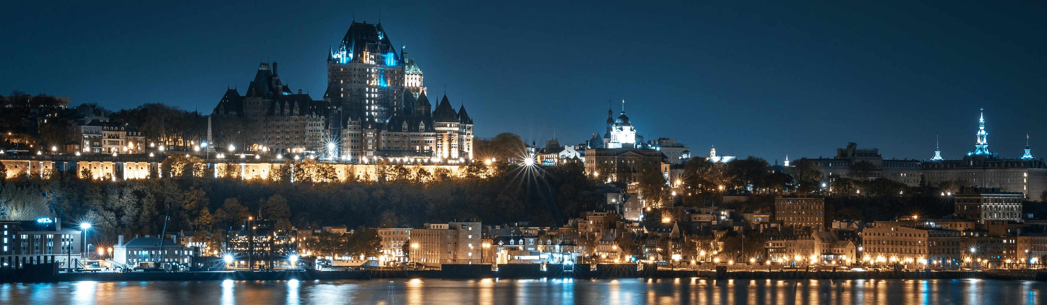A panoramic night view of a historic cityscape with Château Frontenac illuminated atop a hill in Québec City, reflecting in the St. Lawrence River below. 