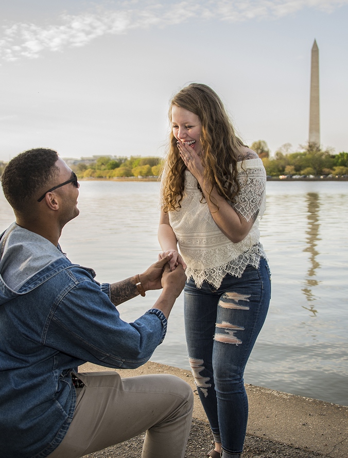 Man proposing to a smiling woman near a body of water with the Washington Monument in the background