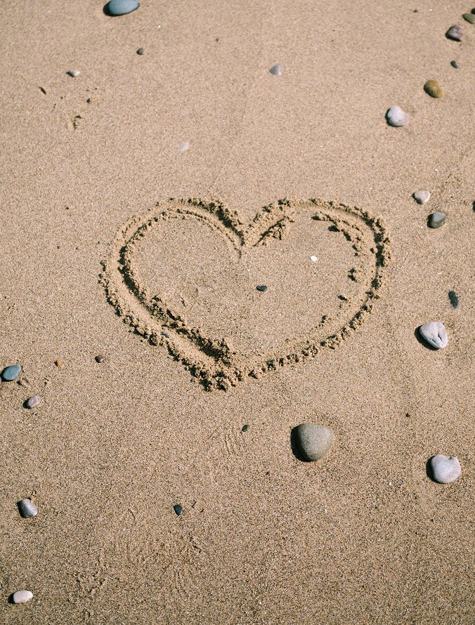 Heart shape drawn in the sand surrounded by scattered pebbles on a beach.
