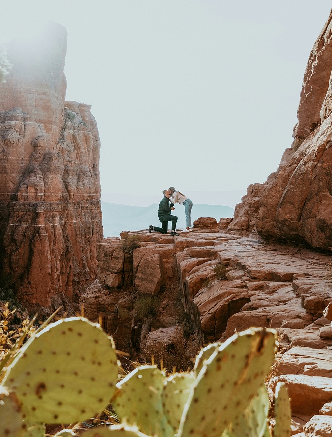 Man proposing to a woman on a scenic cliffside surrounded by red rock formations.