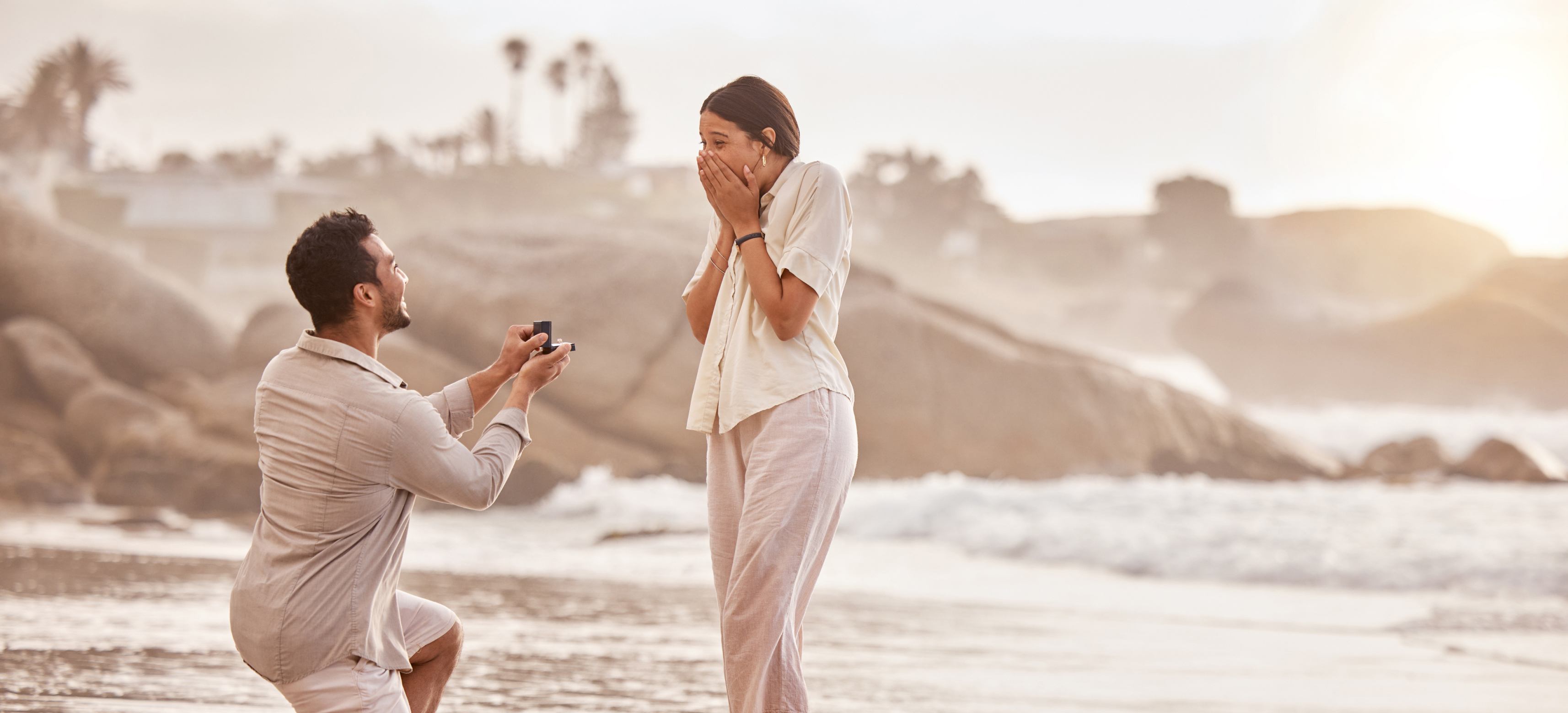 Man proposing to a surprised woman on a beach at sunset, holding an open ring box while kneeling on one knee